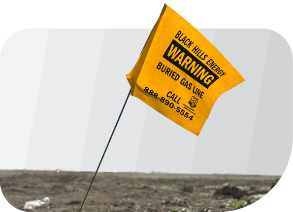 Yellow flag in dirt reads "Black Hills Energy Warning Buried Gas Line Call 888-890-5554," highlighting the presence of underground gas, fiber, or phone lines—call before digging to avoid disrupting essential services.